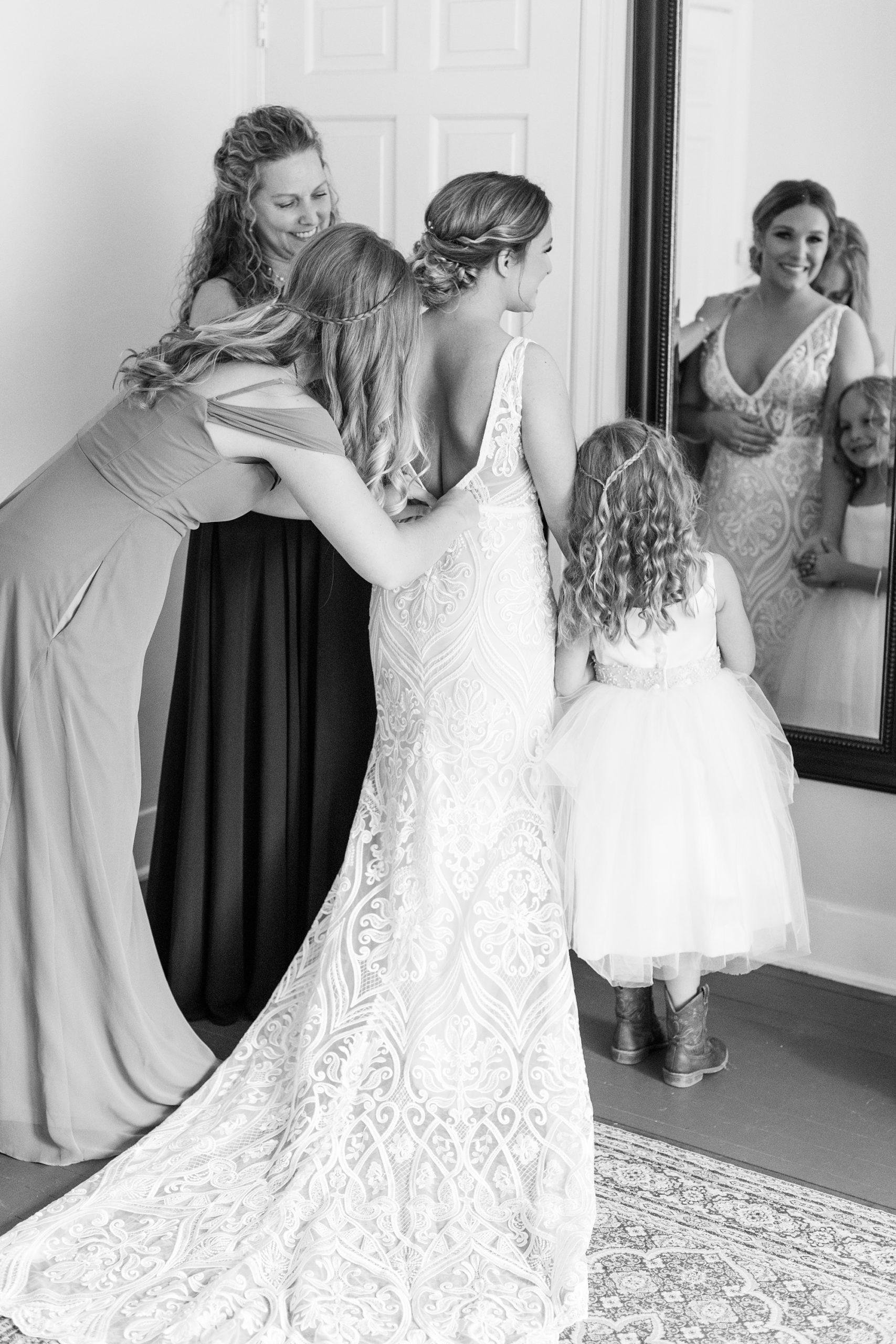 bridesmaid helps bride with gown while daughter watches on